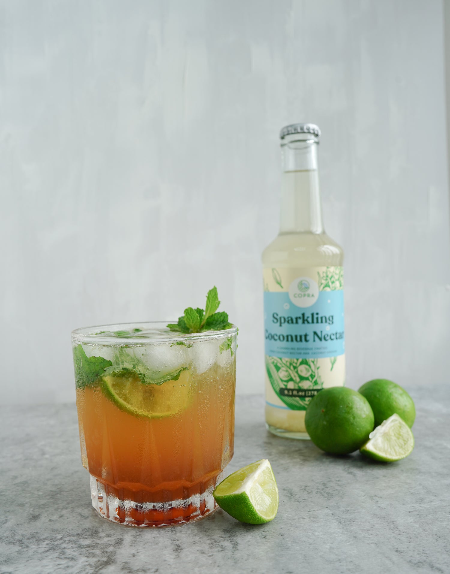 Copra Sparkling Coconut Nectar bottle next to a a glass containing a mixed drink with limes and mint.