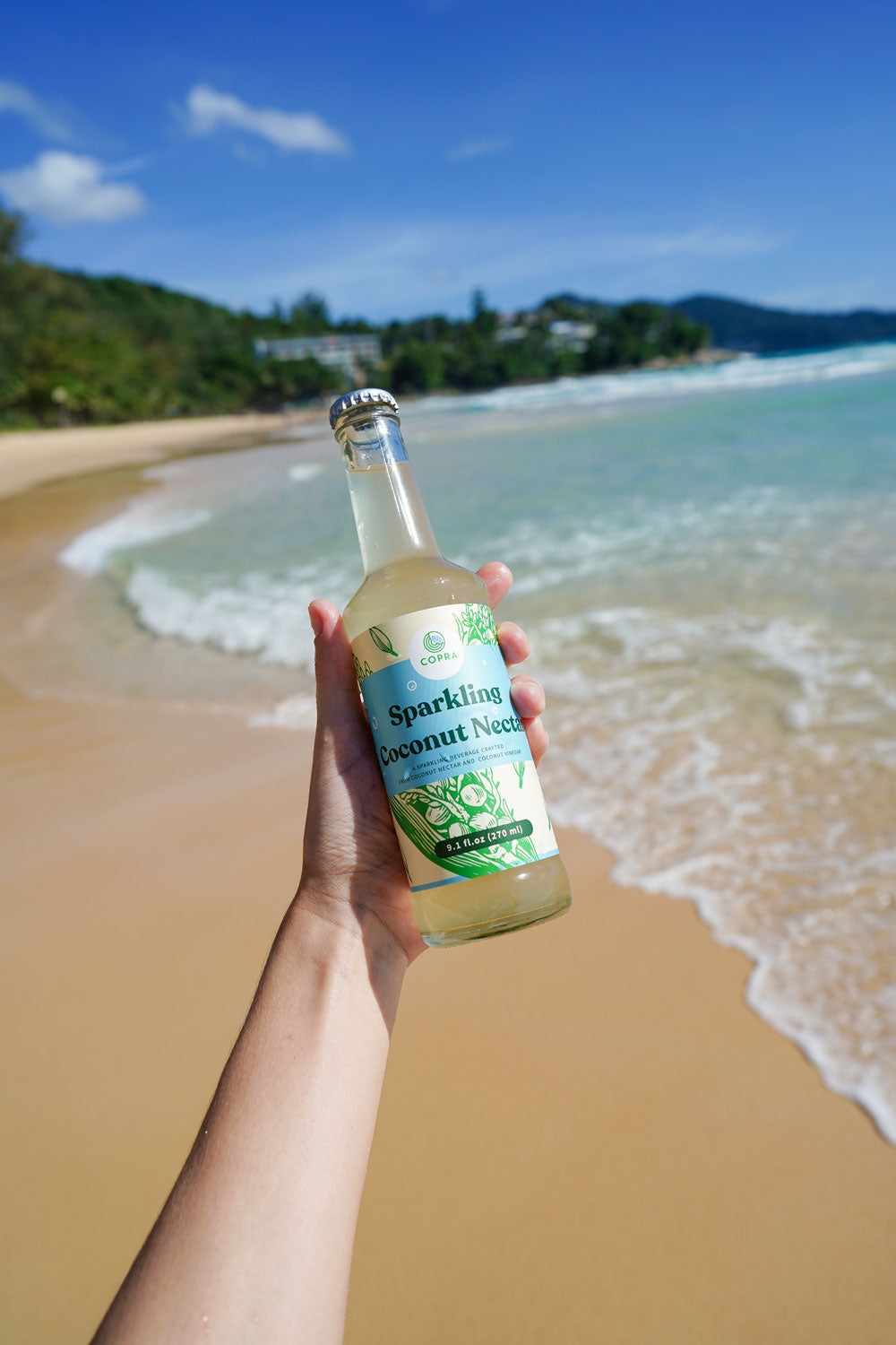 Copra Coconut Sparkling Coconut Nectar held up in front of a beautiful beach scene in Thailand.